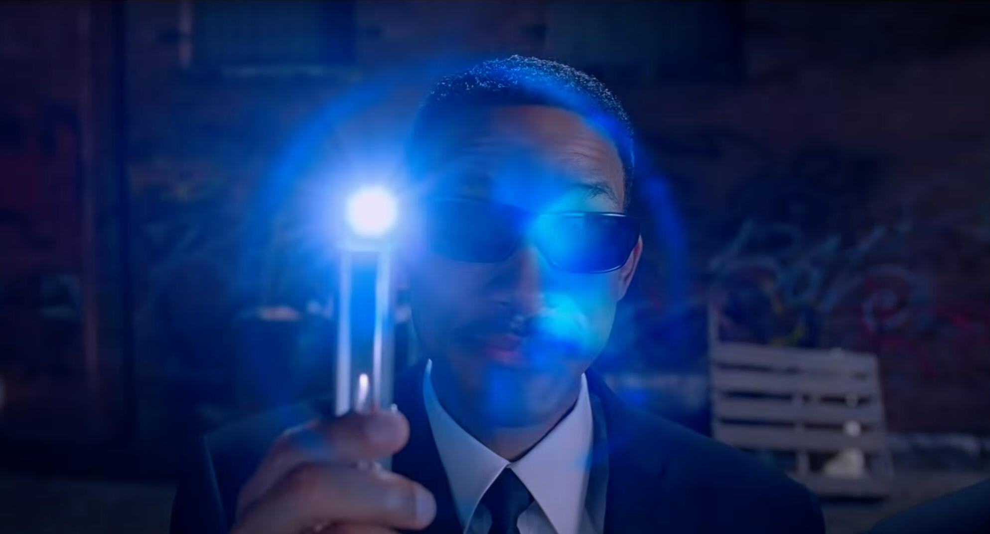 The actor Will Smith dressed as a Men In Black Agent holding a neuralyzer that is emitting a blinding blue light from the top of the device